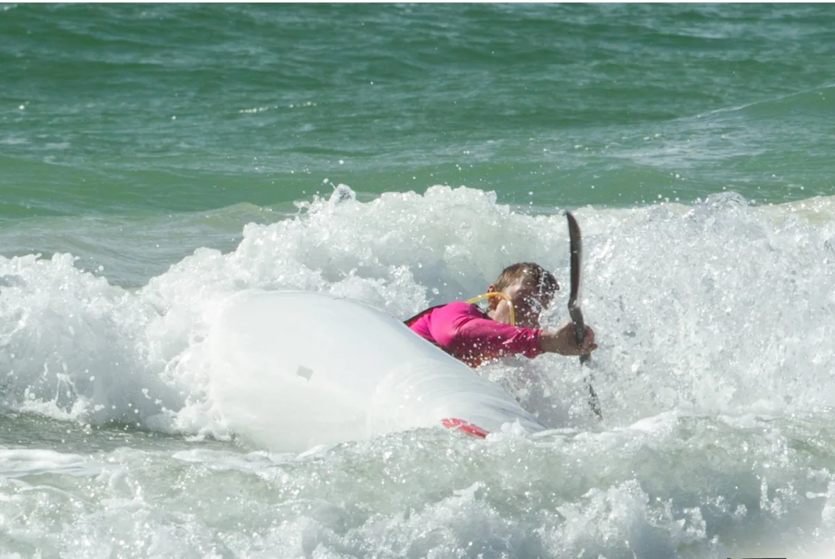 Kristina mid-stroke through breaking surf in a pink rashie, ocean spray flying &mdash; competitive ocean paddling at a race start